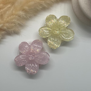 Peut inclure: Deux supports de téléphone en forme de fleurs scintillantes. L'un est jaune clair, l'autre rose. Les deux ont une base transparente et sont décorés de paillettes scintillantes. Les pétales de fleurs sont détaillés avec des nervures subtiles.