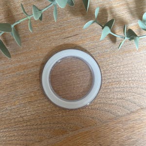 Peut inclure: Un objet circulaire transparent avec un bord blanc, possiblement un couvercle ou un élément décoratif. Il est posé sur une surface en bois brun clair, avec des brins de feuilles vertes en arrière-plan.