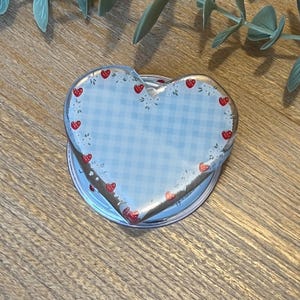 Peut inclure: Un dessous de verre en acrylique transparent en forme de cœur avec un motif vichy bleu clair. Le dessous de verre est décoré de petites fraises rouges sur le bord. Le dessous de verre repose sur une base ronde de couleur argent.