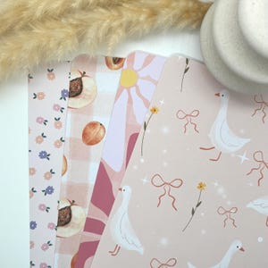 Peut inclure: Une collection de quatre feuilles de papier décoratives avec divers motifs. Les motifs comprennent de petites fleurs, des moitiés de pêches sur un fond à carreaux, une marguerite rétro et des oies blanches avec des nœuds. La palette de couleurs est rose clair, pêche et blanc.