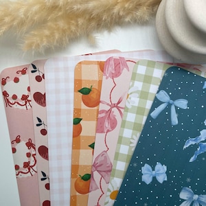 Peut inclure: Six feuilles de papier à motifs différents. Les motifs comprennent un vichy rose et blanc, un vichy vert et blanc, une feuille bleue à pois blancs et nœuds bleus, une feuille rose à nœuds blancs et fraises, une feuille orange à vichy blanc et oranges, et une feuille rose à cerises et un gâteau.