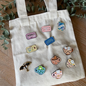 Peut inclure: Une collection d'épingles émaillées sur un sac fourre-tout en toile. Les épingles présentent divers motifs, dont une chauve-souris, une grenouille dans une tasse à thé et des billets avec les textes "Spooky Book Club" et "Romance Book Club".
