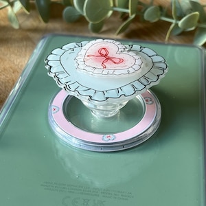 Peut inclure: Un support de téléphone transparent avec un motif de cœur rose et blanc. Le cœur est orné d'un nœud rouge et entouré d'un bord festonné. Le support est fixé à une base circulaire rose et blanche.