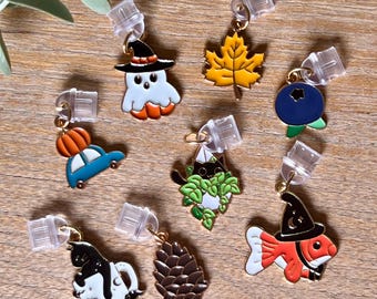 May include: A collection of colorful enamel charms with gold-tone findings. The charms include a ghost in a witch hat, a maple leaf, a blueberry, a car with a pumpkin, a cat in leaves, a cat in a teacup, a pinecone, and a fish in a witch hat.
