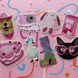 May include: A collection of seven stickers featuring a golden retriever in a pink cowboy hat, a pink camera, a light pink plush rabbit, a pink juice carton labeled Creative Juices, a heart-shaped cake, a goose with blue flowers, and a black cat.