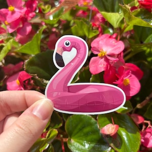 May include: A pink flamingo-shaped sticker with a black beak and eye is held in front of a background of pink flowers and green leaves. The sticker is a cartoon illustration of a flamingo pool float.