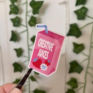May include: A pink sticker in the shape of a juice box with the words "CREATIVE JUICES" and strawberry illustrations. The sticker also includes the text "10g sugar". A blue straw is visible.