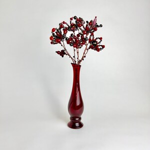 May include: A red glass vase with a beaded flower arrangement in shades of red and black. The vase has a rounded base and a narrow neck.