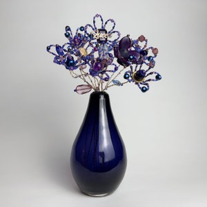 May include: A blue glass vase with a beaded flower arrangement in shades of purple, blue, and gold. The flower arrangement features a gold charm that says "Queen".
