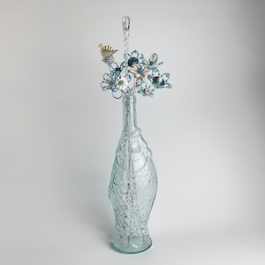 Puede incluir: Un jarrón de vidrio transparente con forma de pez con un arreglo floral de cuentas en su interior. Las flores están hechas de cuentas azules y blancas y hay un pequeño pájaro posado en una rama.