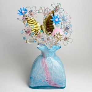 Pode incluir: Um vaso de vidro azul e branco com uma borda ondulada contém um buquê de flores e borboletas de contas. As flores são feitas de contas azuis e rosas, e as borboletas são douradas.