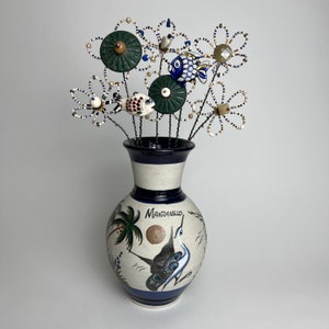 Puede incluir: Un jarrón de cerámica blanca con detalles pintados en azul y verde, que presenta un diseño de pájaro y la palabra "Manzanillo". El jarrón está lleno de un ramo de flores de cuentas de colores.