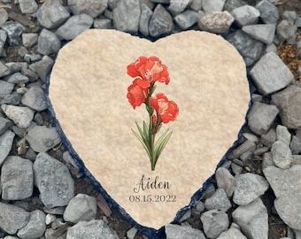 May include: Heart-shaped stone with a painted design of red flowers and green leaves. The stone has the name "Aiden" and the date "08.15.2022" written on it.