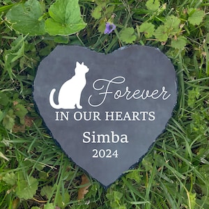May include: A heart-shaped, dark gray slate memorial stone with a white cat silhouette and the words "Forever In Our Hearts Simba 2024". The stone is set in green grass.