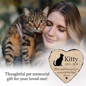 May include: A person holds a tabby cat, with a heart-shaped memorial stone in the foreground. The stone features a black cat silhouette, the name "Kitty", dates 2013-2024, and the inscription "If love could have saved you you would have lived forever."