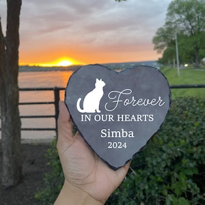 May include: A heart-shaped, gray slate pet memorial with a white cat silhouette and the words "Forever In Our Hearts" and "Simba 2024". The memorial is held up against a sunset backdrop.