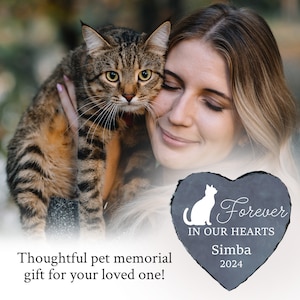 May include: A tabby cat is held by a person, with a heart-shaped pet memorial stone in the foreground. The stone is gray with a white cat silhouette, the words "Forever In Our Hearts," and the name "Simba" with the year "2024."