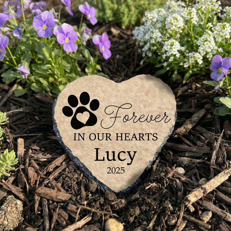 Heart-shaped natural beige stone pet memorial placed in a garden with purple flowers. It features a black paw print and the text "Forever IN OUR HEARTS," personalized with "Lucy 2025".