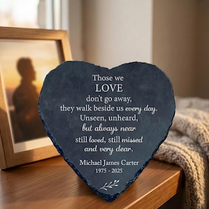 May include: A heart-shaped memorial stone with engraved text. The dark gray stone features the words "Those we LOVE" and a personalized message, along with a name and dates. A framed photo is visible in the background.