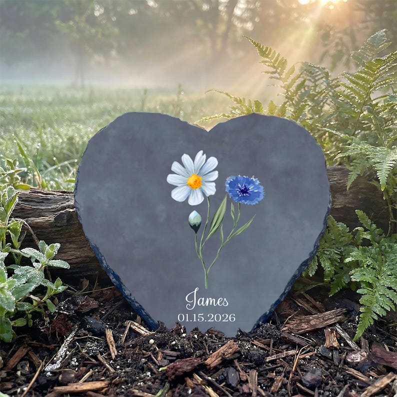 Heart-shaped slate memorial stone placed in a garden with tulips. It features a watercolor painting of a white daisy and blue cornflower, personalized with the name "James" and date "01.15.2026."