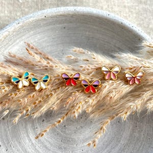 Könnte beinhalten: Sechs Schmetterlings-Ohrstecker mit goldfarbenen Rahmen und Emaille-Flügeln in Weiß, Blau, Rot, Lila und Rosa. Die Ohrringe sind auf trockenem, weizenfarbenem Gras in einer grauen, kreisförmigen Schale angeordnet.