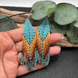 Op de afbeelding: Een paar turquoise, bruine en gouden kralen oorbellen met een franje ontwerp.