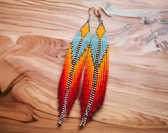 Op de afbeelding: Een paar lange, met kralen bezette oorbellen met een levendig kleurenschema. De oorbellen hebben een combinatie van turquoise, gele, oranje en rode kralen, gerangschikt in een geometrisch patroon. Het franjedesign voegt beweging en flair toe aan deze statement oorbellen.