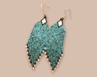 Op de afbeelding: Teal kralen oorbellen met een franjedesign. De oorbellen hebben goudkleurige haken en kleine goudkleurige kralen aan de onderkant van de franje. De bovenkant van de oorbellen heeft een zwarte rand met een witte ruitvorm.