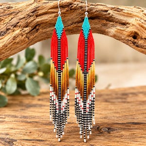 May include: A pair of long, dangling earrings crafted from tiny seed beads. The design features a triangular top in turquoise, transitioning into a vertical gradient of red, orange, yellow, and black, ending in a fringe of black and white beads.