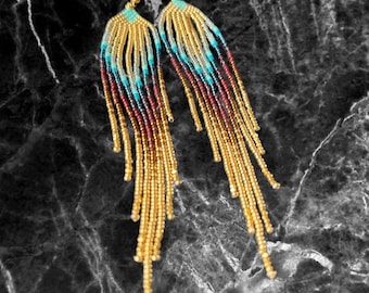 Op de afbeelding: Lange, geperde oorbellen met een gouden, turquoise en bruine kleurstelling. De oorbellen zijn gemaakt van kleine, ronde kralen en hebben een kwastachtig ontwerp.