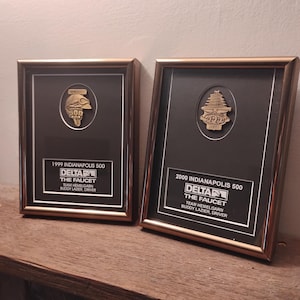 Może przedstawiać: Dwie oprawione tablice pamiątkowe. Każda z nich posiada złoty emblemat, czarne tło i złotą ramę. Tablice upamiętniają wyścigi Indianapolis 500 z 1999 i 2000 roku, z tekstem identyfikującym zesp&oacute;ł i kierowcę.