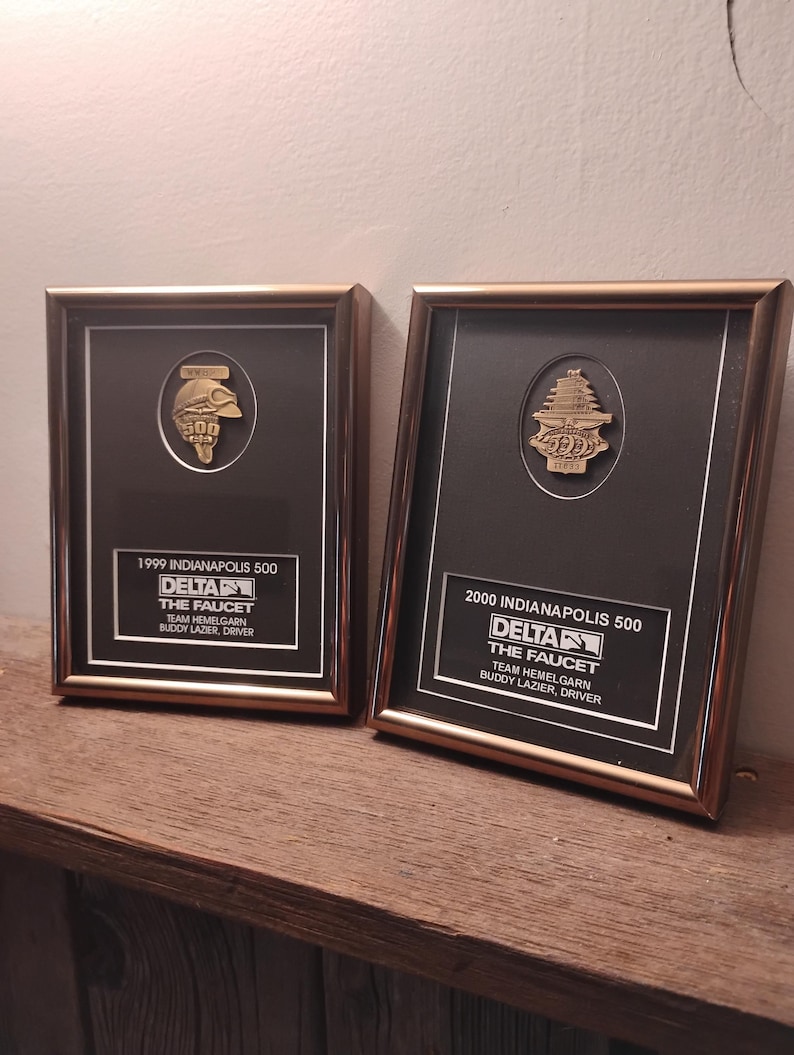 Może przedstawiać: Dwie oprawione tablice pamiątkowe. Każda z nich posiada złoty emblemat, czarne tło i złotą ramę. Tablice upamiętniają wyścigi Indianapolis 500 z 1999 i 2000 roku, z tekstem identyfikującym zesp&oacute;ł i kierowcę.