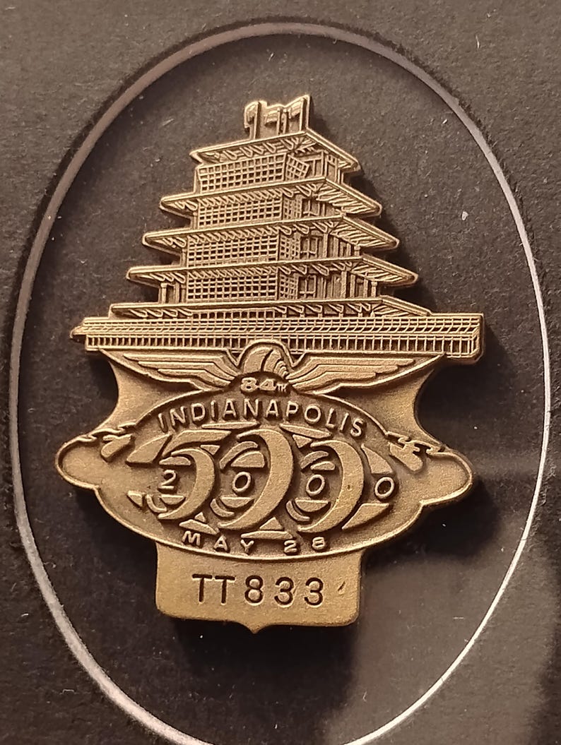 Może przedstawiać: Brązowa przypinka z wielopoziomowym projektem pagody nad emblematem w kształcie tarczy. Na emblemacie widnieje napis "84th Indianapolis 500 May 26" z wyraźnie widoczną liczbą "2000". Przypinka ma r&oacute;wnież napis "TT833".