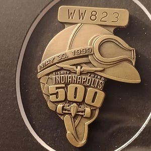 Może przedstawiać: Brązowa przypinka upamiętniająca Indianapolis 500. Przypinka przedstawia kask wyścigowy z goglami, napis "MAY 30, 1999" oraz "EIGHTY THIRD INDIANAPOLIS 500". Przypinka zawiera r&oacute;wnież napis "WW 823".