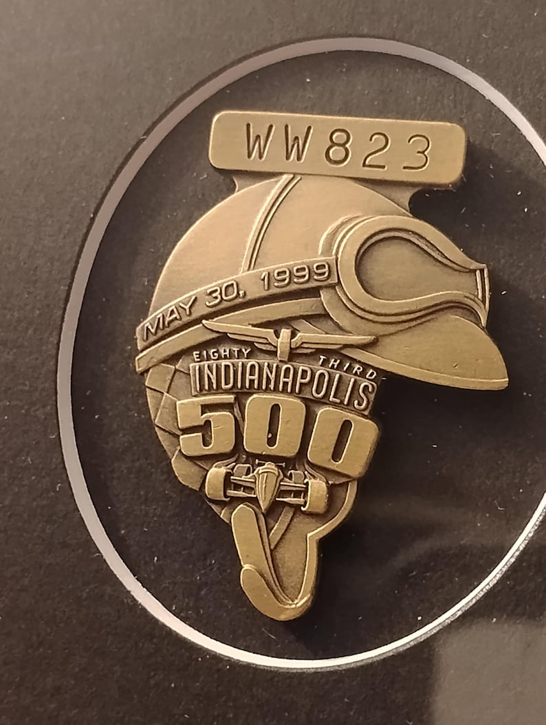 Może przedstawiać: Brązowa przypinka upamiętniająca Indianapolis 500. Przypinka przedstawia kask wyścigowy z goglami, napis "MAY 30, 1999" oraz "EIGHTY THIRD INDIANAPOLIS 500". Przypinka zawiera r&oacute;wnież napis "WW 823".