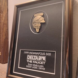 Może przedstawiać: Oprawiona tablica pamiątkowa z Indianapolis 500 z 1999 roku. Czarne tło zawiera złoty emblemat i tekst "1999 Indianapolis 500, Delta, The Faucet, Team Hemelgarn, Buddy Lazier, Driver."
