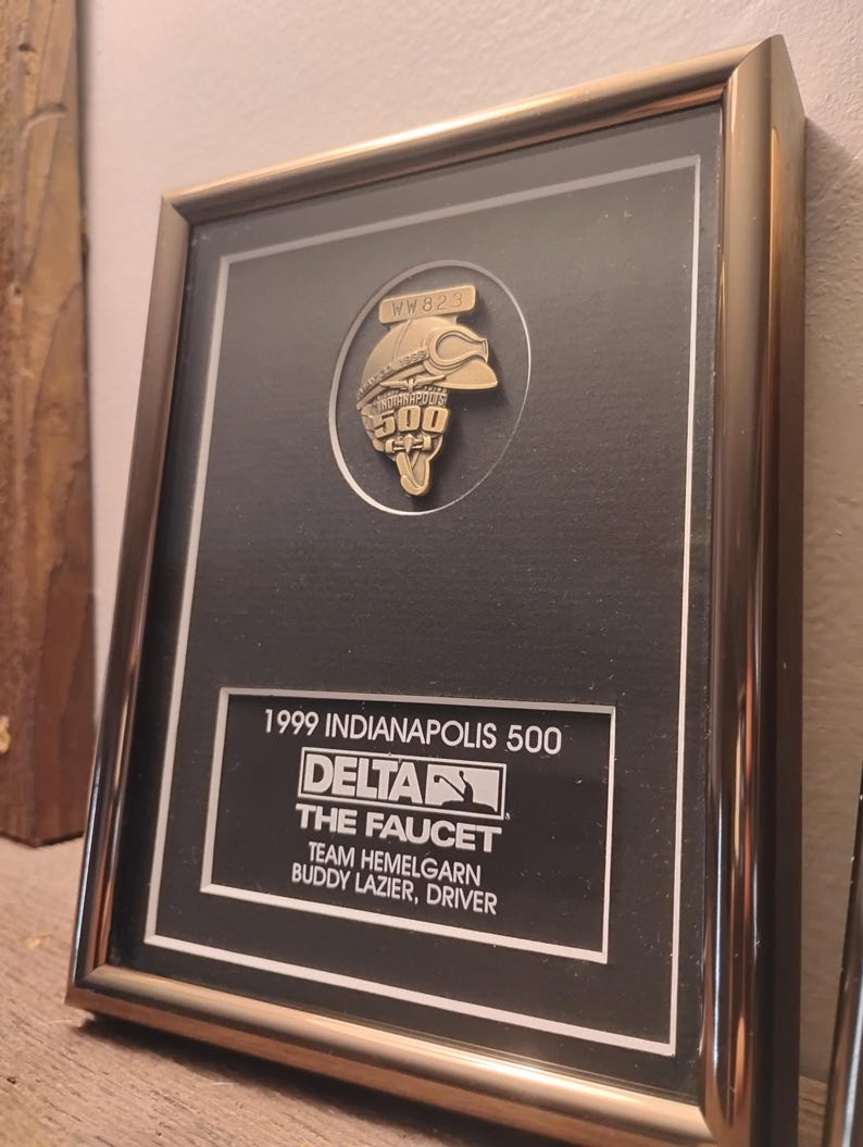 Może przedstawiać: Oprawiona tablica pamiątkowa z Indianapolis 500 z 1999 roku. Czarne tło zawiera złoty emblemat i tekst "1999 Indianapolis 500, Delta, The Faucet, Team Hemelgarn, Buddy Lazier, Driver."