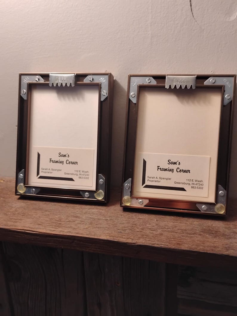 Może przedstawiać: Dwie prostokątne brązowe ramy do zdjęć z metalowymi narożnikami i wieszakami z ząbkami. Każda rama ma białe wnętrze i etykietę z napisem "Sam's Framing Corner" z danymi kontaktowymi.