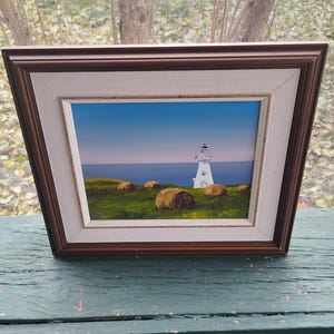 May include: Framed painting of a white lighthouse on a grassy hill with hay bales, under a blue sky. The artwork is in a brown and white frame, displayed on a teal surface.
