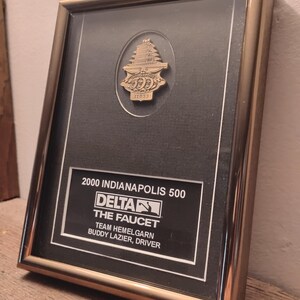 Może przedstawiać: Oprawiona tablica pamiątkowa z wyścigu Indianapolis 500 z 2000 roku. Złota rama otacza czarne tło z tekstem i złotą przypinką z motywem pagody. Tekst zawiera "2000 Indianapolis 500" i "Delta".