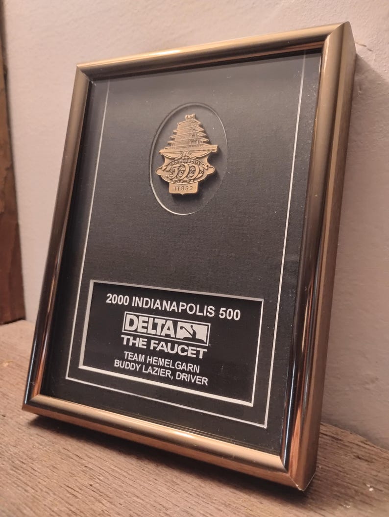 Może przedstawiać: Oprawiona tablica pamiątkowa z wyścigu Indianapolis 500 z 2000 roku. Złota rama otacza czarne tło z tekstem i złotą przypinką z motywem pagody. Tekst zawiera "2000 Indianapolis 500" i "Delta".