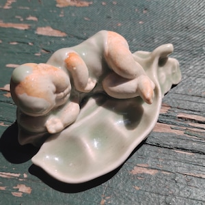 May include: A ceramic figurine of a baby lying on a leaf-shaped dish. The baby has a light green body with peach-colored accents on the head and limbs. The leaf dish is also light green, with a glossy finish.