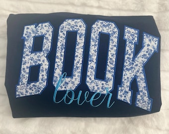May include: A navy blue sweatshirt with the word "BOOK" in white with a blue floral pattern and the word "lover" in blue cursive lettering.