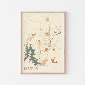 May include: Framed map print of Branson, Missouri, USA. The map features a beige background with brown, orange, and teal accents. The city's name is printed in a brown font at the bottom. The map is enclosed in a light brown wooden frame.