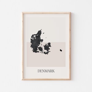 May include: A framed print showing a minimalist map of Denmark in black against a beige backdrop. The word "DENMARK" is printed in a simple font at the bottom. The print is encased in a light wood frame.