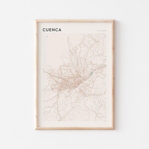 May include: Framed print of Cuenca, Ecuador, featuring a detailed map in shades of beige and brown. The city name is printed in black at the top left. The print is encased in a light-colored wooden frame.