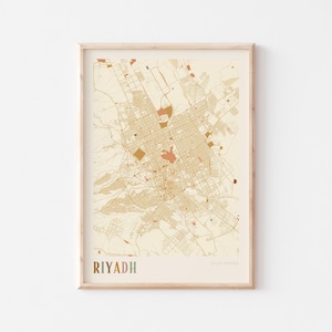 Op de afbeelding: Een ingelijste kaartprint van Riyad, Saoedi-Arabië, in een neutraal kleurenpalet. De stadskaart heeft een gedetailleerd stratennet met beige, bruine en oranje tinten. Het woord "RIYADH" staat onderaan in kleurrijke letters.