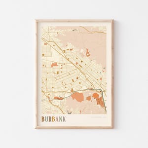May include: A framed map print of Burbank, California, USA. The map features a detailed street layout in shades of beige, brown, and orange, with the city name at the bottom. The print is encased in a light-colored wooden frame.