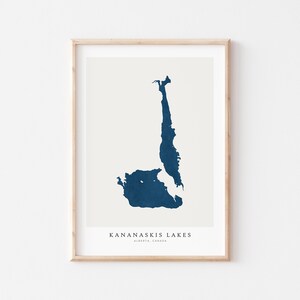 Pode incluir: Uma impressão emoldurada com um mapa estilizado dos lagos Kananaskis, Alberta, Canadá, em um tom azul escuro sobre um fundo creme. A impressão está emoldurada em uma moldura de madeira clara, com o texto "KANANASKIS LAKES" e "ALBERTA, CANADA" na parte inferior.