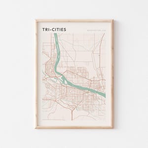 May include: A framed map print titled TRI-CITIES WASHINGTON, USA. The map features a light pink background with thin brown lines representing roads and a prominent light green shape depicting the river flowing through the city area.